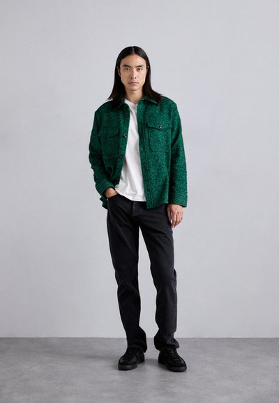 Veste chemise texturée verte avec deux poches poitrine, portée sur un t-shirt blanc, accompagnée de jeans noirs et de chaussures noires. Design simple.