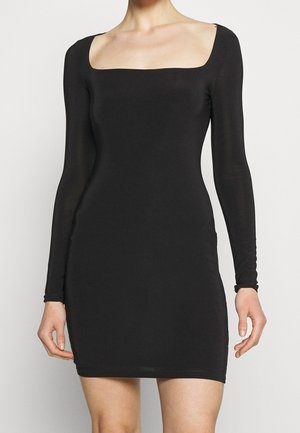 Woman wearing a fitted black long-sleeve mini dress with a wide square neckline, standing against a plain light background.