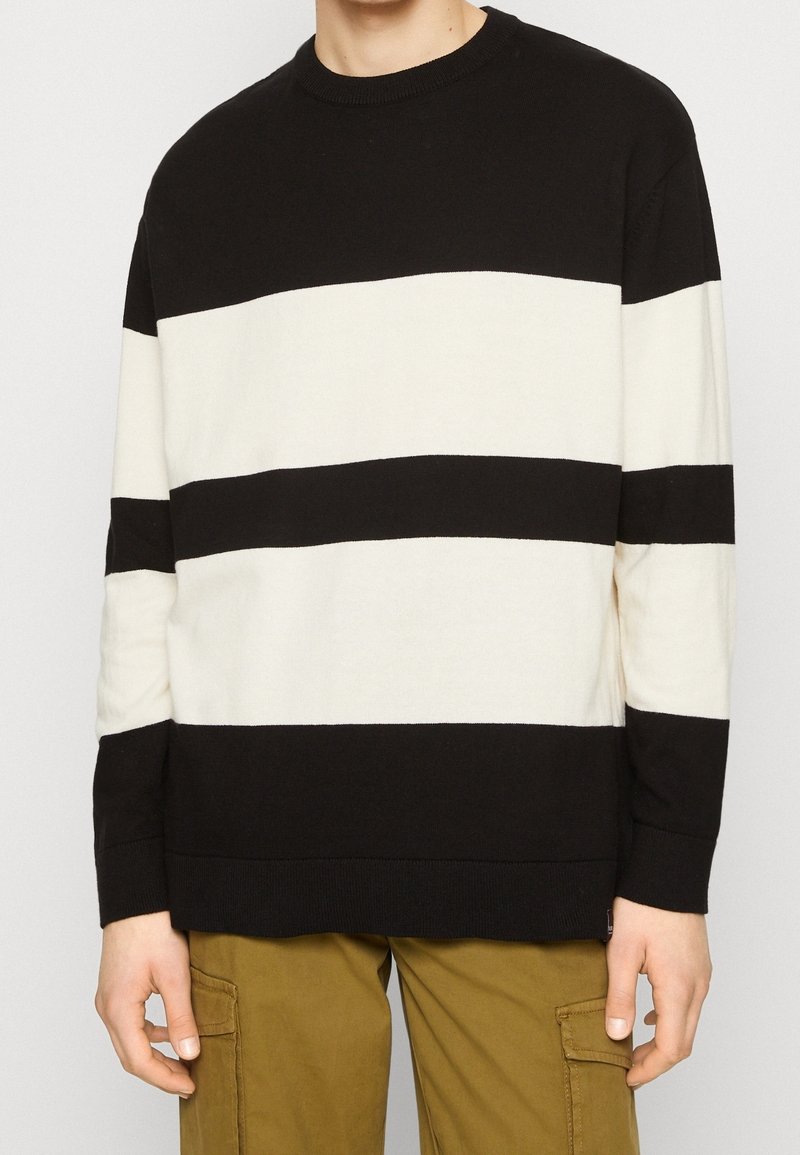 Jersey de rayas negras y crema con cuello redondo, mangas largas y puños y dobladillo acanalados. La tela parece suave y texturizada.