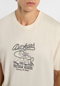 Krémové bavlněné tričko s vintage černou grafikou kovboje a textem "Dickies Uniform Maker Strong As Steel" níže.