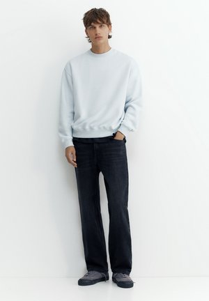 Jeune homme debout, portant un sweat-shirt gris clair, un jean noir et des baskets grises, sur fond blanc uni.
