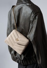 Borsa a tracolla beige trapuntata con hardware dorato, presenta una tracolla a catena e un design elegante, indossata sopra una giacca di pelle scura.
