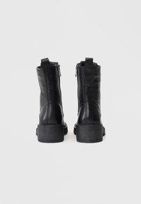 Botas de tobillo de cuero negro con cierre de cremallera, acabado texturizado y tacón de goma apilado. El detalle trasero presenta una costura decorativa.