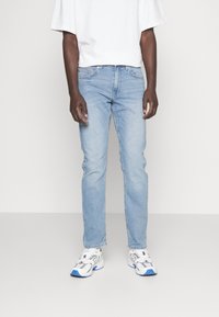 Light blue denim jeans with a slim fit, featuring five pockets and a standard waistband. Worn with a white t-shirt and athletic shoes.