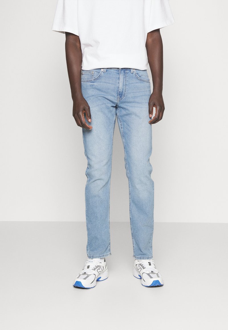 Light blue denim jeans with a slim fit, featuring five pockets and a standard waistband. Worn with a white t-shirt and athletic shoes.