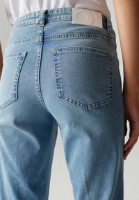 Jeans in denim blu chiaro con vita alta, tasche posteriori e una toppa bianca con logo in vita. Tessuto liscio con sfumature delicate.