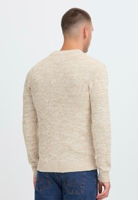 Mann med kort hår som vender bort fra kameraet, iført en beige melert genser og blå jeans foran en enkel hvit bakgrunn.