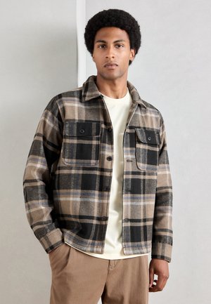 Checked flannel jacket in tan, black, and blue. Features two chest pockets with button closures and a spread collar. Soft fabric texture.