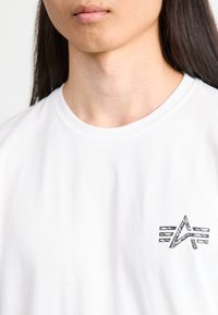 Weißes Baumwoll-T-Shirt mit einem kleinen schwarzen Grafiklogo auf der linken Brust, kurzen Ärmeln und einem klassischen Rundhalsausschnitt.
