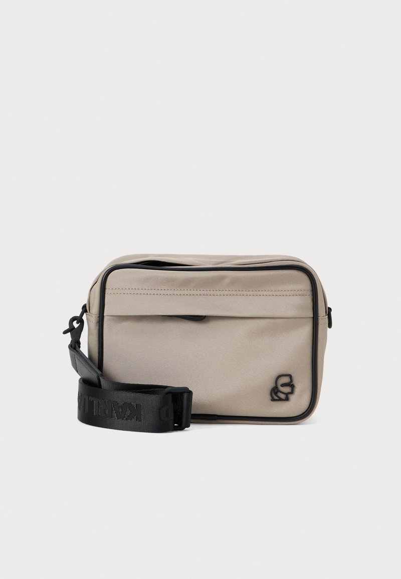 Beżowa prostokątna torba crossbody z czarnym zamkiem błyskawicznym, szczegółami logo i regulowanym paskiem. Gładki materiał tekstylny, minimalistyczny design.