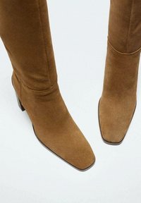 Bottines en suède marron montantes jusqu'au genou avec un bout carré et un talon bloc. Le matériau a une texture douce et une finition lisse.