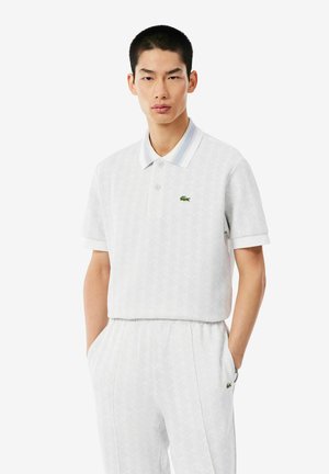 Hombre con polo blanco de manga corta y pantalones blancos a juego con las manos en los bolsillos sobre un fondo liso.