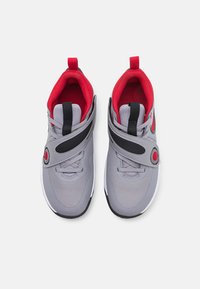 Zapatillas deportivas grises con acentos rojos, con parte superior de malla, correa ajustable y suela texturizada. Incluyen un detalle circular en la correa.