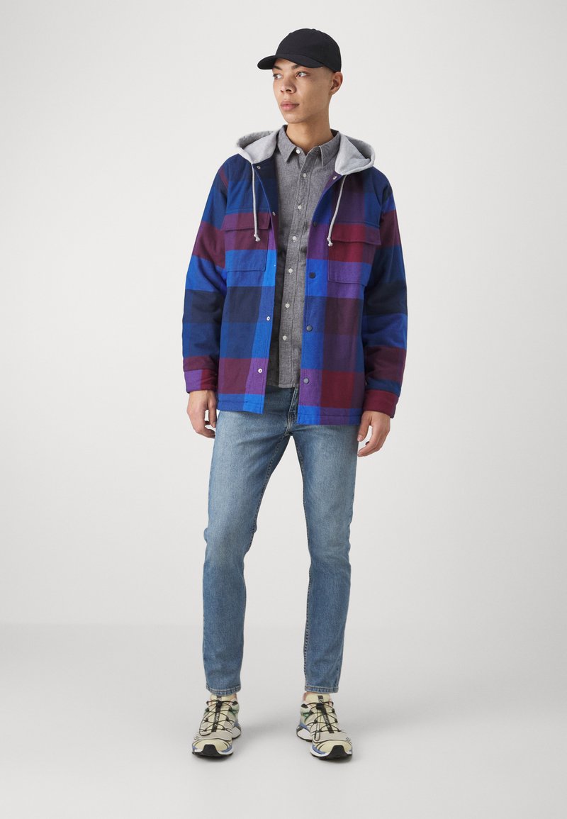 Gestapeltes Outfit mit einer blau-burgunderfarbenen Karo-Jacke mit grauem Kapuzenstück, getragen über einem grauen Hemd mit Knöpfen und hellen Jeans, abgerundet mit bunten Sneakern.