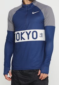 Man wearing a blue and gray long-sleeve athletic top with a white band labeled "TOKYO" and a small Nike logo on the chest.