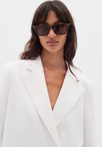 Bela platnena blazer z globokim V izrezom, z odrezanimi klapami in gladko teksturo. Model nosi oversized rjave mačje očala.