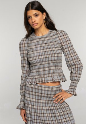 Mujer con cabello largo y oscuro que lleva un top fruncido de cuadros marrón y azul y una falda a juego, posando con una mano en la cadera sobre un fondo liso.