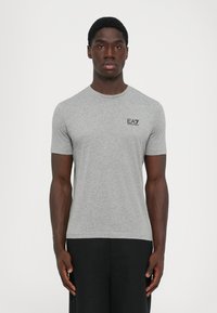Mann iført en tettsittende grå kortermet t-skjorte med "EA7 Emporio Armani"-logo på brystet og svarte bukser, som står mot en ensfarget bakgrunn.