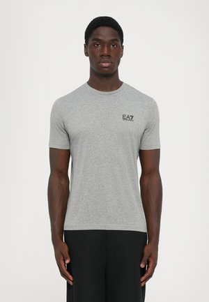 Man wearing a fitted grey short-sleeve t-shirt with "EA7 Emporio Armani" logo on chest and black pants, standing against a plain background.