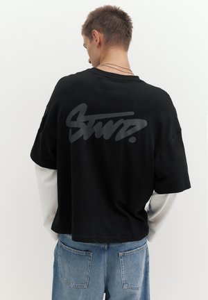 Ung mand med kort hår iført lagdelte sorte og hvide skjorter og blå jeans, vendt væk med stiliseret tekst på bagsiden af den sorte skjorte.