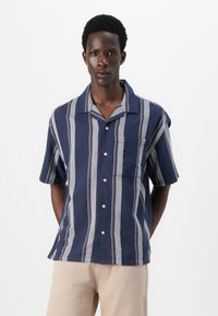 Chemise à manches courtes marine avec des rayures verticales blanches et rouges. Dispose d'un devant à boutons et d'une poche poitrine. Fabriquée en tissu léger.