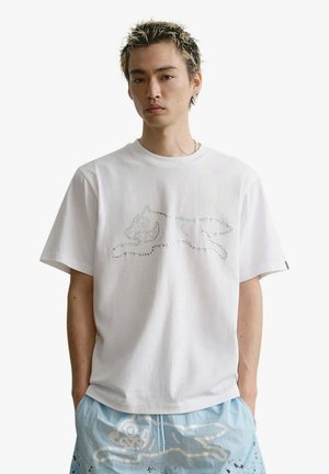 Giovane uomo con capelli biondi a punte che indossa una maglietta bianca con il contorno di una lepre in corsa realizzato con strass e pantaloncini azzurri chiari con stampa di lepre.