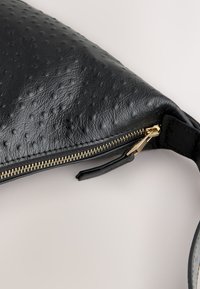 Black textured leather pouch with gold zipper and black leather pull tab on a light surface.