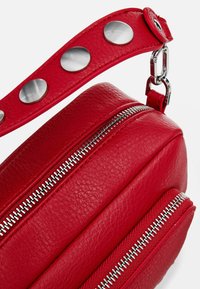 Runde rote Lederhandtasche mit zwei silbernen Reißverschlüssen und einem Trageriemen, der mit metallischen Nieten verziert ist. Die Oberfläche hat eine strukturierte Verarbeitung.