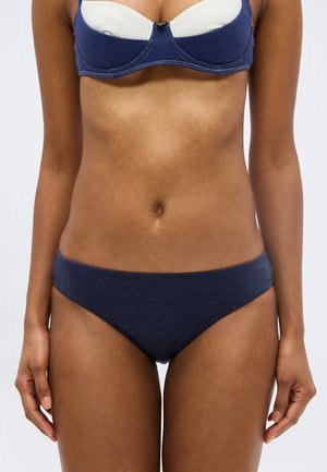 Žena v tmavomodrých bikinách so spodným dielom a bielym s tmavomodrými detailmi na hornom diele, stojaca s rukami voľne pozdĺž tela.