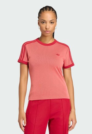 Kvinde med flettet hår iført en rød Adidas t-shirt med tre striber på ærmerne og matchende røde bukser mod en ensfarvet baggrund.