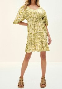 Robe à manches courtes, verte à motifs avec une taille cintrée, un ourlet à volants et des motifs floraux. Portée avec des sandales marron à lanières.