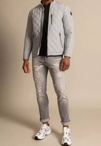 Giacca trapuntata grigia, t-shirt nera, jeans chiari graffiati e sneakers bianche con dettagli neri. Presenta tasche con zip e maniche arrotolate.