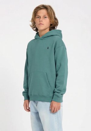 Junge mit schulterlangen Haaren, der einen türkisfarbenen Kapuzenpullover und hellblaue Jeans trägt, steht vor einem schlichten weißen Hintergrund.