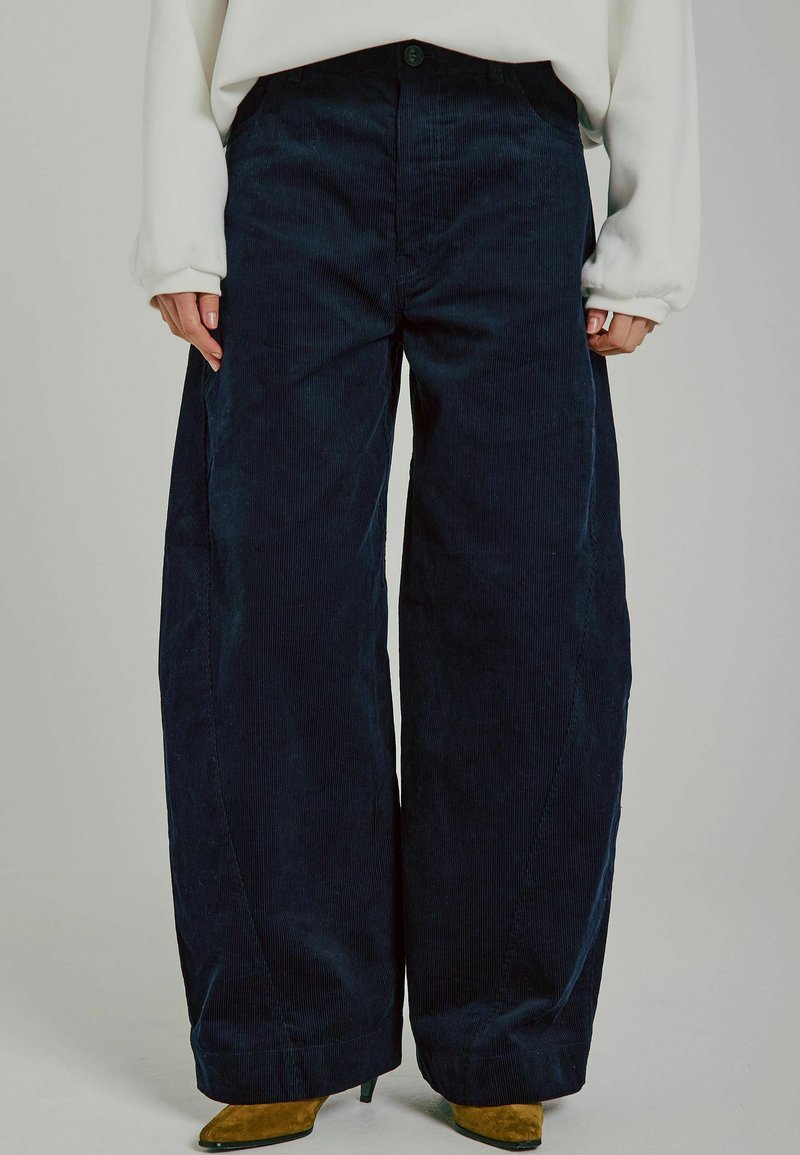Dunkelblaue Cordhosen mit weitem Bein und hoher Taille, vorn mit Knopfverschluss und zwei Seitentaschen. Glatte Textur mit vertikalen Rippungen.