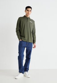 Dolga rokavna olivna polo majica s ovratnikom in detajlom logotipa, v kombinaciji z modrimi kavbojkami in belimi supergami. Minimalističen dizajn.