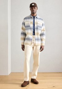 Leichte, gemusterte Bluse in Blau, Gelb und Weiß mit zwei Brusttaschen, über einem dunkelblauen Oberteil getragen und kombiniert mit cremfarbenen Hosen und braunen Stiefeln.