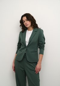 Groene blazer met een getailleerd ontwerp, met een gelapt revers en twee voorzakken, gecombineerd met een witte t-shirt en bijpassende broek.