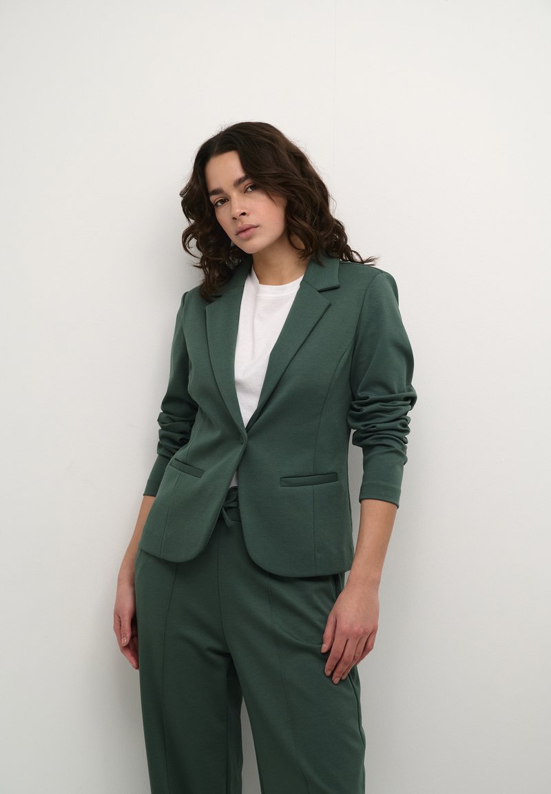 Groene blazer met een getailleerd ontwerp, met een gelapt revers en twee voorzakken, gecombineerd met een witte t-shirt en bijpassende broek.