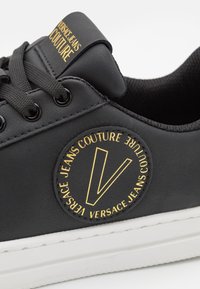 Black sneaker made of synthetic material featuring a round logo patch with gold text, black laces, and a white rubber sole.