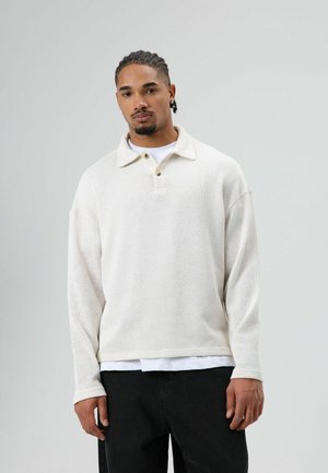 Witte, gestructureerde pullover met een polokraag, twee knopen en lange mouwen. Gedragen over een wit shirt en gecombineerd met een zwarte broek.