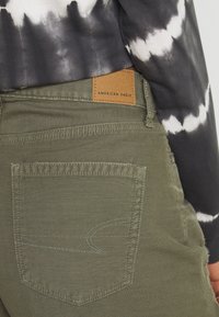 Pantalones de algodón verde oliva con un bolsillo trasero texturizado, que cuenta con costuras blancas y una etiqueta de cuero marrón que dice "American Eagle".