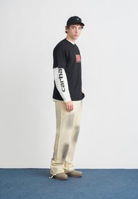 Camiseta gráfica negra combinada con una camisa de manga larga blanca, pantalones beige con un patrón desvaído y zuecos grises. La gorra negra completa el conjunto.