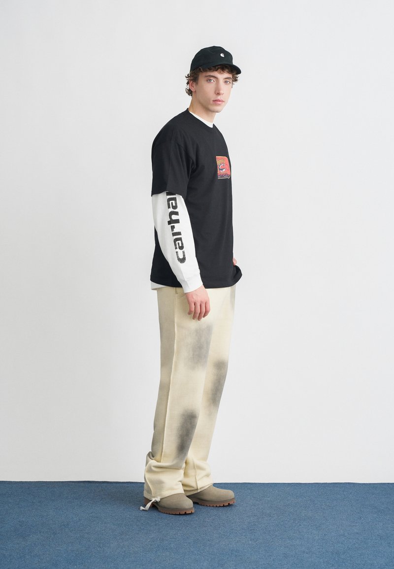 Zwart grafisch T-shirt gedragen over een wit shirt met lange mouwen, beige broek met een vervaald patroon en grijze klompen. Een zwarte pet maakt de outfit compleet.