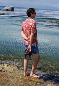 Man met een bloes met bloemenprint en korte broek met patroon staat op blote voeten op een rotsachtige kust, houdt sandalen vast en kijkt uit over de kalme zee.