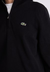 Pulover tricotat negru cu fermoar pe un sfert, având un logo brodat cu crocodil verde și alb pe partea stângă a pieptului.