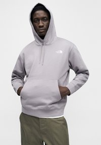 Γκρι φούτερ με κουκούλα και μπροστινή τσέπη, με μικρό λευκό λογότυπο. Το ύφασμα φαίνεται μαλακό, με χαλαρή εφαρμογή και μακριά μανίκια.