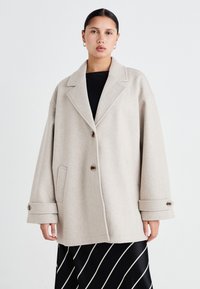 Beige villakangastakki, jossa on viistetty kaulus, nappikiinnitys, sivutaskut ja käännetyt hihansuut. Kangas on pehmeä rakenne; käytetään mustan paidan päällä.