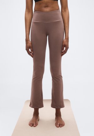 Person stående barfodet på måtte iført højttaljede lilla viddebukser og en sort cropped top, vist fra torso midt til fødder.