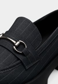 Mocassin gris foncé présentant des rayures verticales, un bout arrondi, une texture semblable au cuir et un accent de boucle métallique sur le dessus.