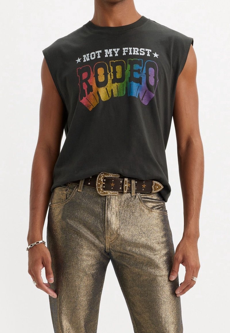 T-shirt sans manches noir avec le texte multicolore "CE N'EST PAS MA PREMIÈRE RODEO", associé à un pantalon doré brillant et une ceinture ornée de clous décoratifs.